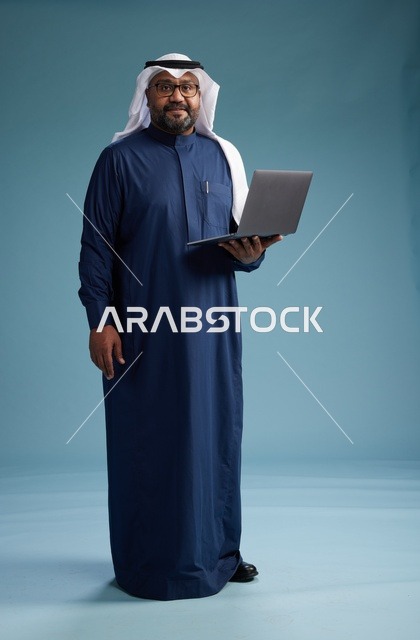 بورتريه لرجل سعودي بثوب أزرق وغترة بيضاء يستخدم الكمبيوتر المحمول, خلفيه زرقاء