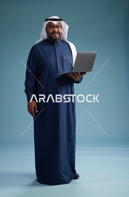 بورتريه لرجل سعودي بثوب أزرق وغترة بيضاء يستخدم الكمبيوتر المحمول, خلفيه زرقاء
