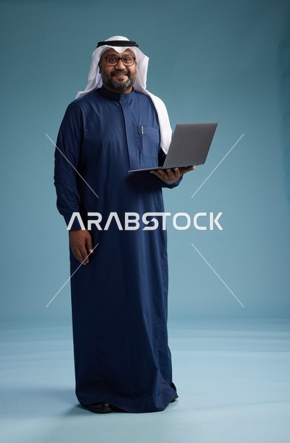 بورتريه لرجل سعودي بثوب أزرق وغترة بيضاء يستخدم الكمبيوتر المحمول, خلفيه زرقاء
