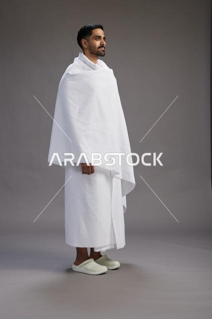 بورتريه لرجل عربي سعودي خليجي يرتدي ملابس الإحرام لأداء مناسك الحج, العباده والتقرب الى الله , الحج والعمره في مكه, الجسم بالطول الكامل , خلفيه رماديه