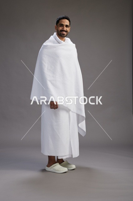 A portrait of an Arab Saudi Gulf man wearing Ihram clothing to perform the rituals of Hajj, worship, and drawing closer to God, Hajj and Umrah in Mecca, full-body, gray background.