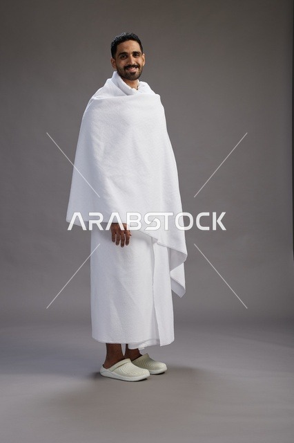 بورتريه لرجل عربي سعودي خليجي يرتدي ملابس الإحرام لأداء مناسك الحج, العباده والتقرب الى الله , الحج والعمره في مكه, الجسم بالطول الكامل , خلفيه رماديه