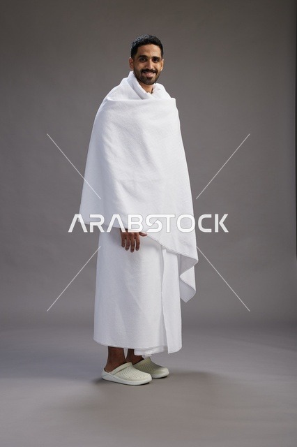 بورتريه لرجل عربي سعودي خليجي يرتدي ملابس الإحرام لأداء مناسك الحج, العباده والتقرب الى الله , الحج والعمره في مكه, الجسم بالطول الكامل , خلفيه رماديه
