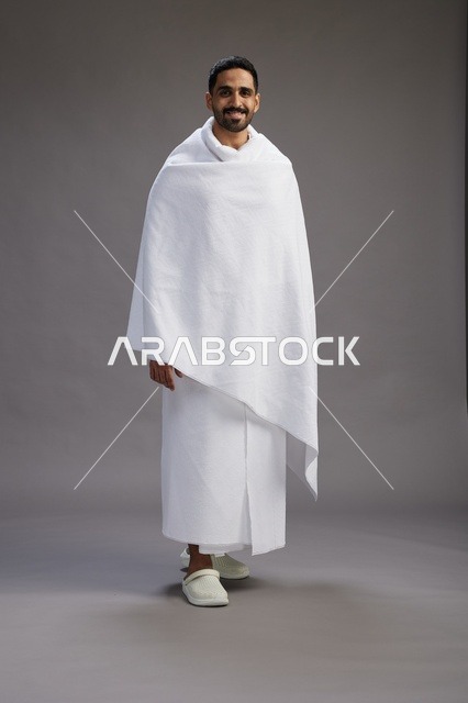 بورتريه لرجل عربي سعودي خليجي يرتدي ملابس الإحرام لأداء مناسك الحج, العباده والتقرب الى الله , الحج والعمره في مكه, الجسم بالطول الكامل , خلفيه رماديه