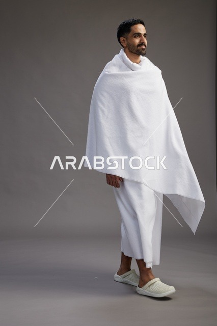 بورتريه لرجل عربي سعودي خليجي يرتدي ملابس الإحرام لأداء مناسك الحج, العباده والتقرب الى الله , الحج والعمره في مكه, الجسم بالطول الكامل , خلفيه رماديه