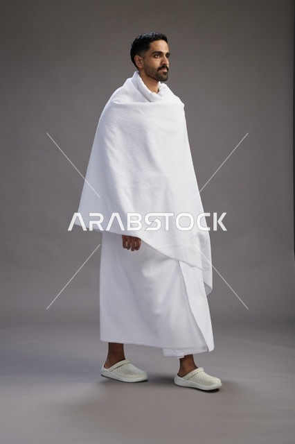 بورتريه لرجل عربي سعودي خليجي يرتدي ملابس الإحرام لأداء مناسك الحج, العباده والتقرب الى الله , الحج والعمره في مكه, الجسم بالطول الكامل , خلفيه رماديه