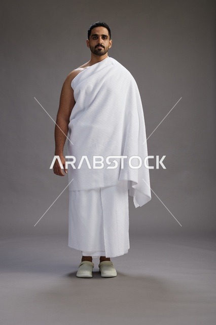 بورتريه لرجل عربي سعودي خليجي يرتدي ملابس الإحرام لأداء مناسك الحج, العباده والتقرب الى الله , الحج والعمره في مكه, الجسم بالطول الكامل , خلفيه رماديه