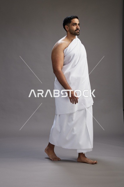 بورتريه لرجل عربي سعودي خليجي يرتدي ملابس الإحرام لأداء مناسك الحج, العباده والتقرب الى الله , الحج والعمره في مكه, الجسم بالطول الكامل , خلفيه رماديه