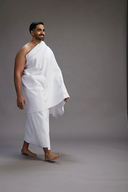 A portrait of an Arab Saudi Gulf man wearing Ihram clothing to perform the rituals of Hajj, worship, and drawing closer to God, Hajj and Umrah in Mecca, full-body, gray background.
