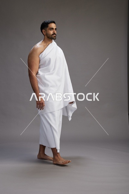 بورتريه لرجل عربي سعودي خليجي يرتدي ملابس الإحرام لأداء مناسك الحج, العباده والتقرب الى الله , الحج والعمره في مكه, الجسم بالطول الكامل , خلفيه رماديه