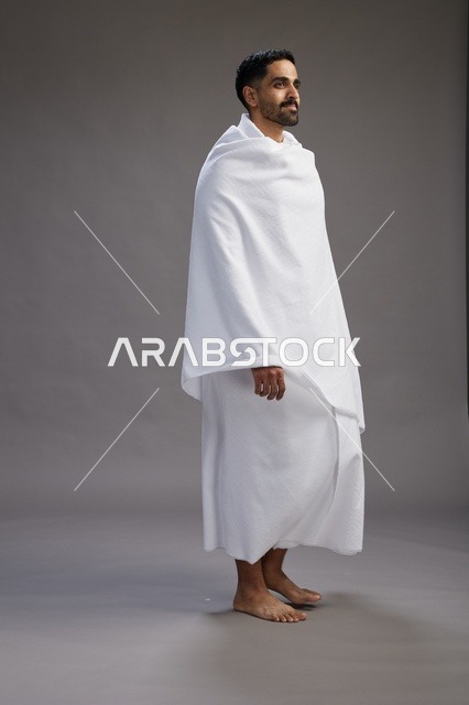A portrait of an Arab Saudi Gulf man wearing Ihram clothing to perform the rituals of Hajj, worship, and drawing closer to God, Hajj and Umrah in Mecca, full-body, gray background.