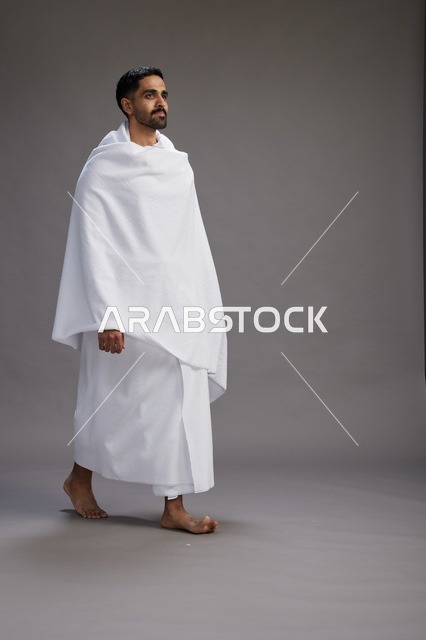 بورتريه لرجل عربي سعودي خليجي يرتدي ملابس الإحرام لأداء مناسك الحج, العباده والتقرب الى الله , الحج والعمره في مكه, الجسم بالطول الكامل , خلفيه رماديه