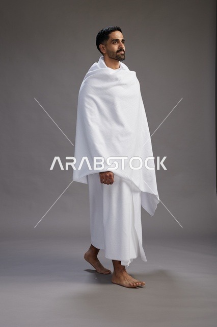 بورتريه لرجل عربي سعودي خليجي يرتدي ملابس الإحرام لأداء مناسك الحج, العباده والتقرب الى الله , الحج والعمره في مكه, الجسم بالطول الكامل , خلفيه رماديه