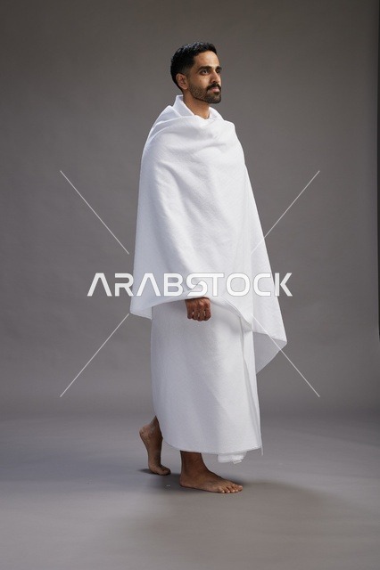 بورتريه لرجل عربي سعودي خليجي يرتدي ملابس الإحرام لأداء مناسك الحج, العباده والتقرب الى الله , الحج والعمره في مكه, الجسم بالطول الكامل , خلفيه رماديه