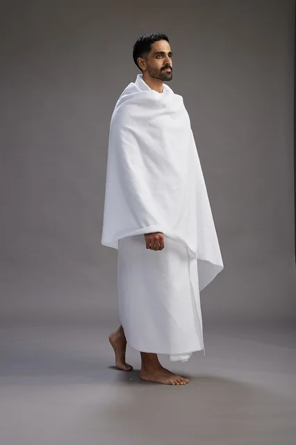 A portrait of an Arab Saudi Gulf man wearing Ihram clothing to perform the rituals of Hajj, worship, and drawing closer to God, Hajj and Umrah in Mecca, full-body, gray background.