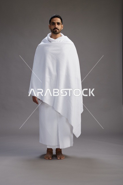 بورتريه لرجل عربي سعودي خليجي يرتدي ملابس الإحرام لأداء مناسك الحج, العباده والتقرب الى الله , الحج والعمره في مكه, الجسم بالطول الكامل , خلفيه رماديه