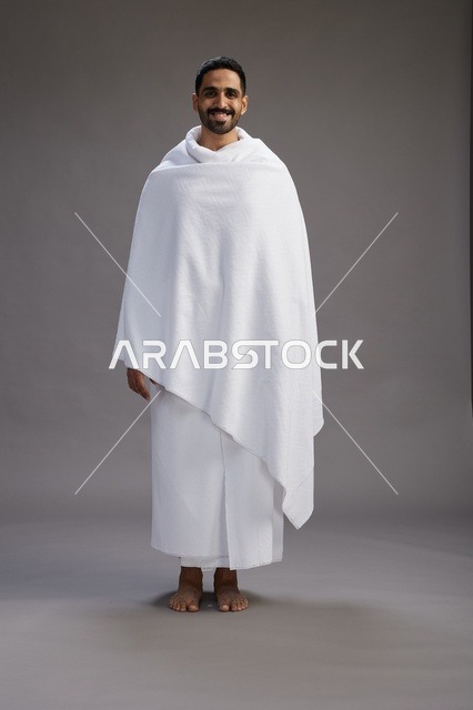 بورتريه لرجل عربي سعودي خليجي يرتدي ملابس الإحرام لأداء مناسك الحج, العباده والتقرب الى الله , الحج والعمره في مكه, الجسم بالطول الكامل , خلفيه رماديه