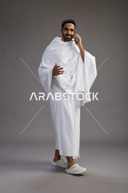 بورتريه لرجل سعودي بملابس الإحرام يجري مكالمة هاتفية عبر الهاتف المحمول, الاستعداد لاداء مناسك الحج والعمره, الجسم بالطول الكامل , خلفيه رماديه