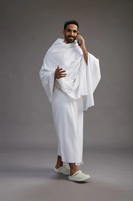 A portrait of a Saudi man in Ihram clothing making a phone call on a mobile phone, preparing to perform the rituals of Hajj and Umrah, full-body shot, gray background.