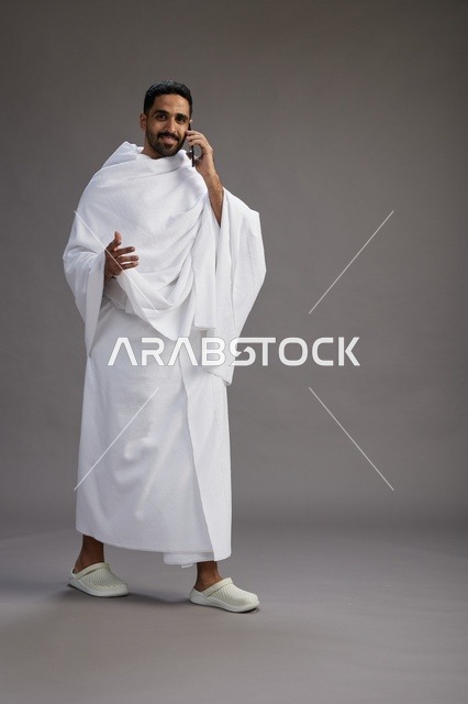 بورتريه لرجل سعودي بملابس الإحرام يجري مكالمة هاتفية عبر الهاتف المحمول, الاستعداد لاداء مناسك الحج والعمره, الجسم بالطول الكامل , خلفيه رماديه