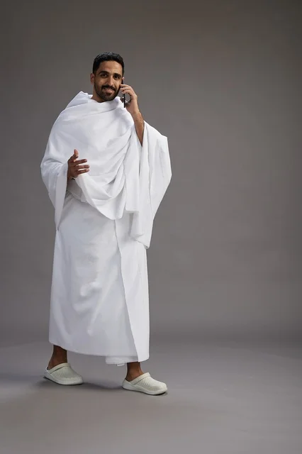 A portrait of a Saudi man in Ihram clothing making a phone call on a mobile phone, preparing to perform the rituals of Hajj and Umrah, full-body shot, gray background.