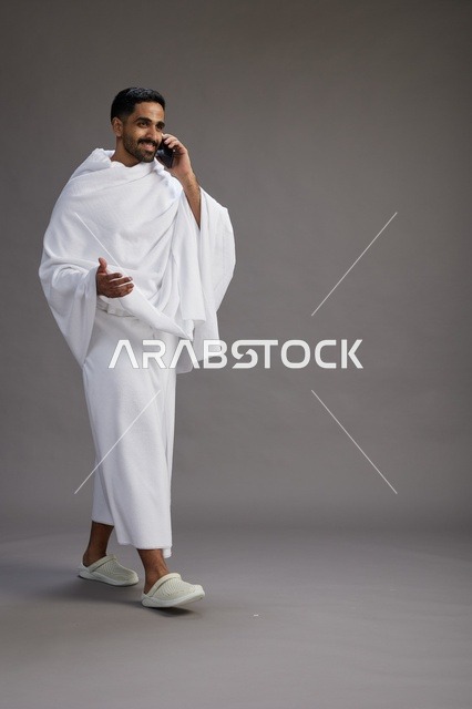 بورتريه لرجل سعودي بملابس الإحرام يجري مكالمة هاتفية عبر الهاتف المحمول, الاستعداد لاداء مناسك الحج والعمره, الجسم بالطول الكامل , خلفيه رماديه