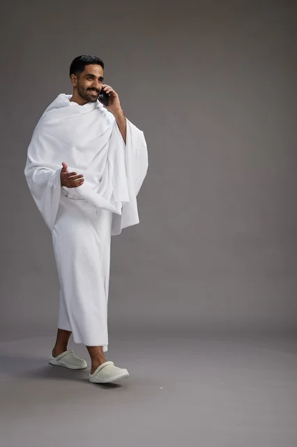 A portrait of a Saudi man in Ihram clothing making a phone call on a mobile phone, preparing to perform the rituals of Hajj and Umrah, full-body shot, gray background.