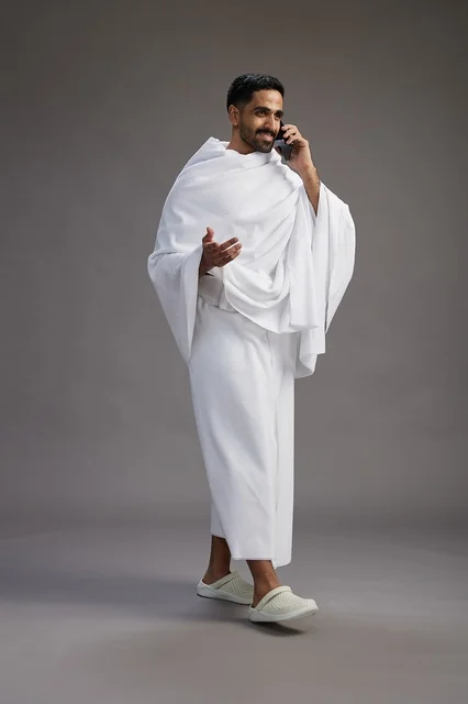 A portrait of a Saudi man in Ihram clothing making a phone call on a mobile phone, preparing to perform the rituals of Hajj and Umrah, full-body shot, gray background. A portrait of a Saudi man in Ihram clothing making a phone call on a mobile phone, preparing to perform the rituals of Hajj and Umrah, full-body shot, gray background.