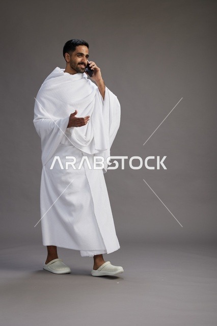 A portrait of a Saudi man in Ihram clothing making a phone call on a mobile phone, preparing to perform the rituals of Hajj and Umrah, full-body shot, gray background.