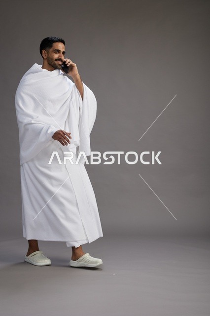 A portrait of a Saudi man in Ihram clothing making a phone call on a mobile phone, preparing to perform the rituals of Hajj and Umrah, full-body shot, gray background.