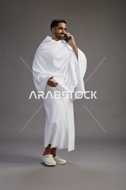 بورتريه لرجل سعودي بملابس الإحرام يجري مكالمة هاتفية عبر الهاتف المحمول, الاستعداد لاداء مناسك الحج والعمره, الجسم بالطول الكامل , خلفيه رماديه
