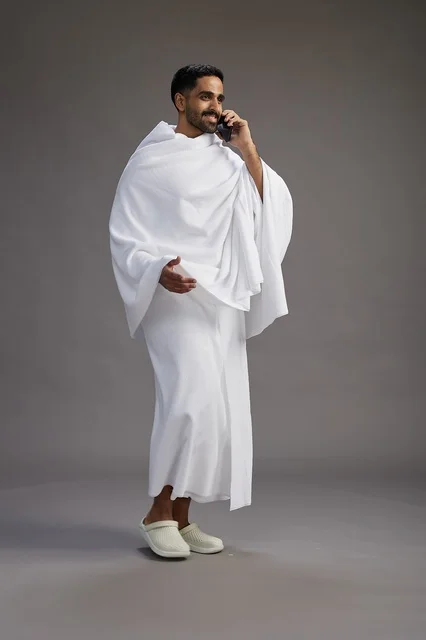 A portrait of a Saudi man in Ihram clothing making a phone call on a mobile phone, preparing to perform the rituals of Hajj and Umrah, full-body shot, gray background.