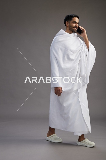 بورتريه لرجل سعودي بملابس الإحرام يجري مكالمة هاتفية عبر الهاتف المحمول, الاستعداد لاداء مناسك الحج والعمره, الجسم بالطول الكامل , خلفيه رماديه