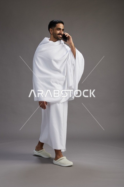 بورتريه لرجل سعودي بملابس الإحرام يجري مكالمة هاتفية عبر الهاتف المحمول, الاستعداد لاداء مناسك الحج والعمره, الجسم بالطول الكامل , خلفيه رماديه