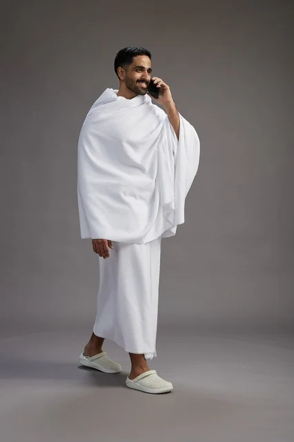 A portrait of a Saudi man in Ihram clothing making a phone call on a mobile phone, preparing to perform the rituals of Hajj and Umrah, full-body shot, gray background.