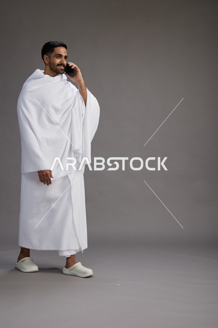 بورتريه لرجل سعودي بملابس الإحرام يجري مكالمة هاتفية عبر الهاتف المحمول, الاستعداد لاداء مناسك الحج والعمره, الجسم بالطول الكامل , خلفيه رماديه