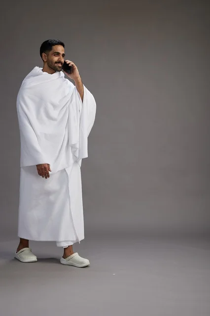 A portrait of a Saudi man in Ihram clothing making a phone call on a mobile phone, preparing to perform the rituals of Hajj and Umrah, full-body shot, gray background. A portrait of a Saudi man in Ihram clothing making a phone call on a mobile phone, preparing to perform the rituals of Hajj and Umrah, full-body shot, gray background.