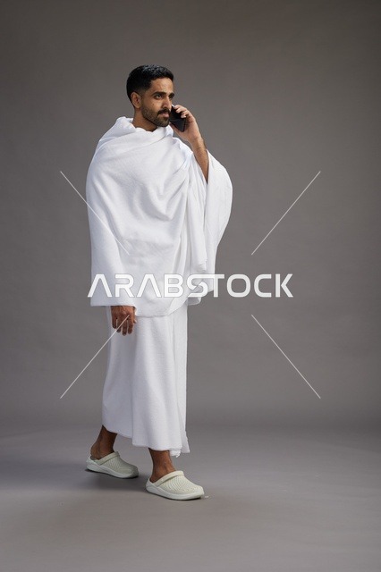 بورتريه لرجل سعودي بملابس الإحرام يجري مكالمة هاتفية عبر الهاتف المحمول, الاستعداد لاداء مناسك الحج والعمره, الجسم بالطول الكامل , خلفيه رماديه