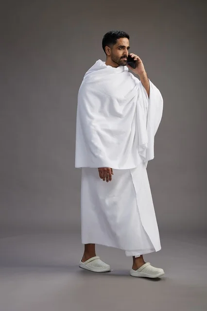 بورتريه لرجل سعودي بملابس الإحرام يجري مكالمة هاتفية عبر الهاتف المحمول, الاستعداد لاداء مناسك الحج والعمره, الجسم بالطول الكامل , خلفيه رماديه