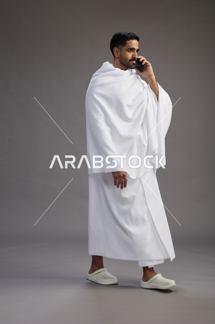 بورتريه لرجل سعودي بملابس الإحرام يجري مكالمة هاتفية عبر الهاتف المحمول, الاستعداد لاداء مناسك الحج والعمره, الجسم بالطول الكامل , خلفيه رماديه