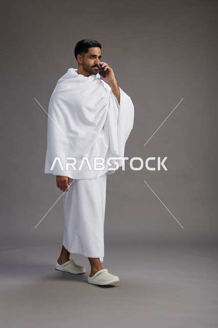 A portrait of a Saudi man in Ihram clothing making a phone call on a mobile phone, preparing to perform the rituals of Hajj and Umrah, full-body shot, gray background.