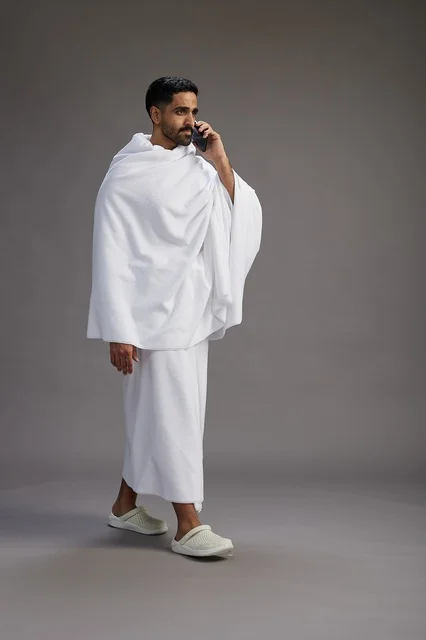 A portrait of a Saudi man in Ihram clothing making a phone call on a mobile phone, preparing to perform the rituals of Hajj and Umrah, full-body shot, gray background.