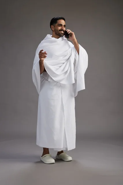 A portrait of a Saudi man in Ihram clothing making a phone call on a mobile phone, preparing to perform the rituals of Hajj and Umrah, full-body shot, gray background.