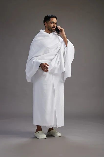 A portrait of a Saudi man in Ihram clothing making a phone call on a mobile phone, preparing to perform the rituals of Hajj and Umrah, full-body shot, gray background.