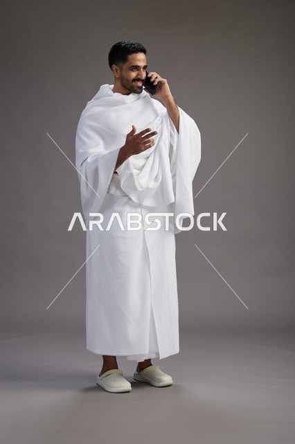 بورتريه لرجل سعودي بملابس الإحرام يجري مكالمة هاتفية عبر الهاتف المحمول, الاستعداد لاداء مناسك الحج والعمره, الجسم بالطول الكامل , خلفيه رماديه