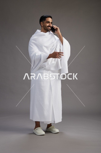بورتريه لرجل سعودي بملابس الإحرام يجري مكالمة هاتفية عبر الهاتف المحمول, الاستعداد لاداء مناسك الحج والعمره, الجسم بالطول الكامل , خلفيه رماديه