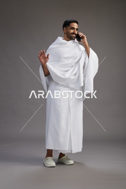 A portrait of a Saudi man in Ihram clothing making a phone call on a mobile phone, preparing to perform the rituals of Hajj and Umrah, full-body shot, gray background.