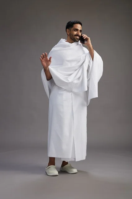 بورتريه لرجل سعودي بملابس الإحرام يجري مكالمة هاتفية عبر الهاتف المحمول, الاستعداد لاداء مناسك الحج والعمره, الجسم بالطول الكامل , خلفيه رماديه
