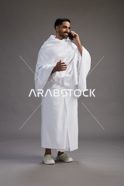 بورتريه لرجل سعودي بملابس الإحرام يجري مكالمة هاتفية عبر الهاتف المحمول, الاستعداد لاداء مناسك الحج والعمره, الجسم بالطول الكامل , خلفيه رماديه