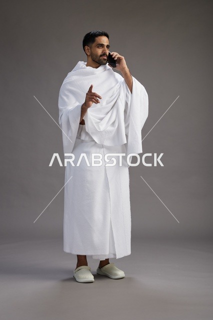 بورتريه لرجل سعودي بملابس الإحرام يجري مكالمة هاتفية عبر الهاتف المحمول, الاستعداد لاداء مناسك الحج والعمره, الجسم بالطول الكامل , خلفيه رماديه
